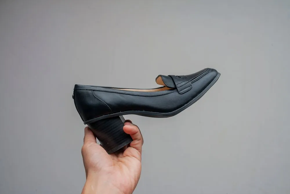 Mano sosteniendo un mocasín negro de taco bajo sobre fondo gris liso