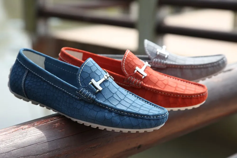 Tres mocasines de cuero grabado en azul, naranja y gris alineados sobre una baranda de madera